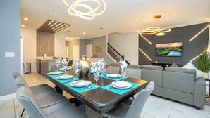 A modern dining area with a long table set for four, a grey sofa, and a staircase in the background. - Private Pool - Themed Rooms - Near Disney 4239 (Kissimmee)