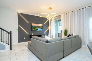 En moderne stue med en grå hjørnesofa, et fladskærms-tv monteret på en mørk væg og en trappe med sorte gelændere.
