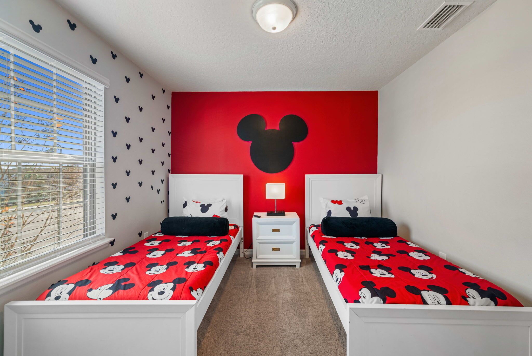 A room with two beds, each with a Mickey Mouse themed bedspread, a red accent wall with a Mickey Mouse head, and a window with blinds.