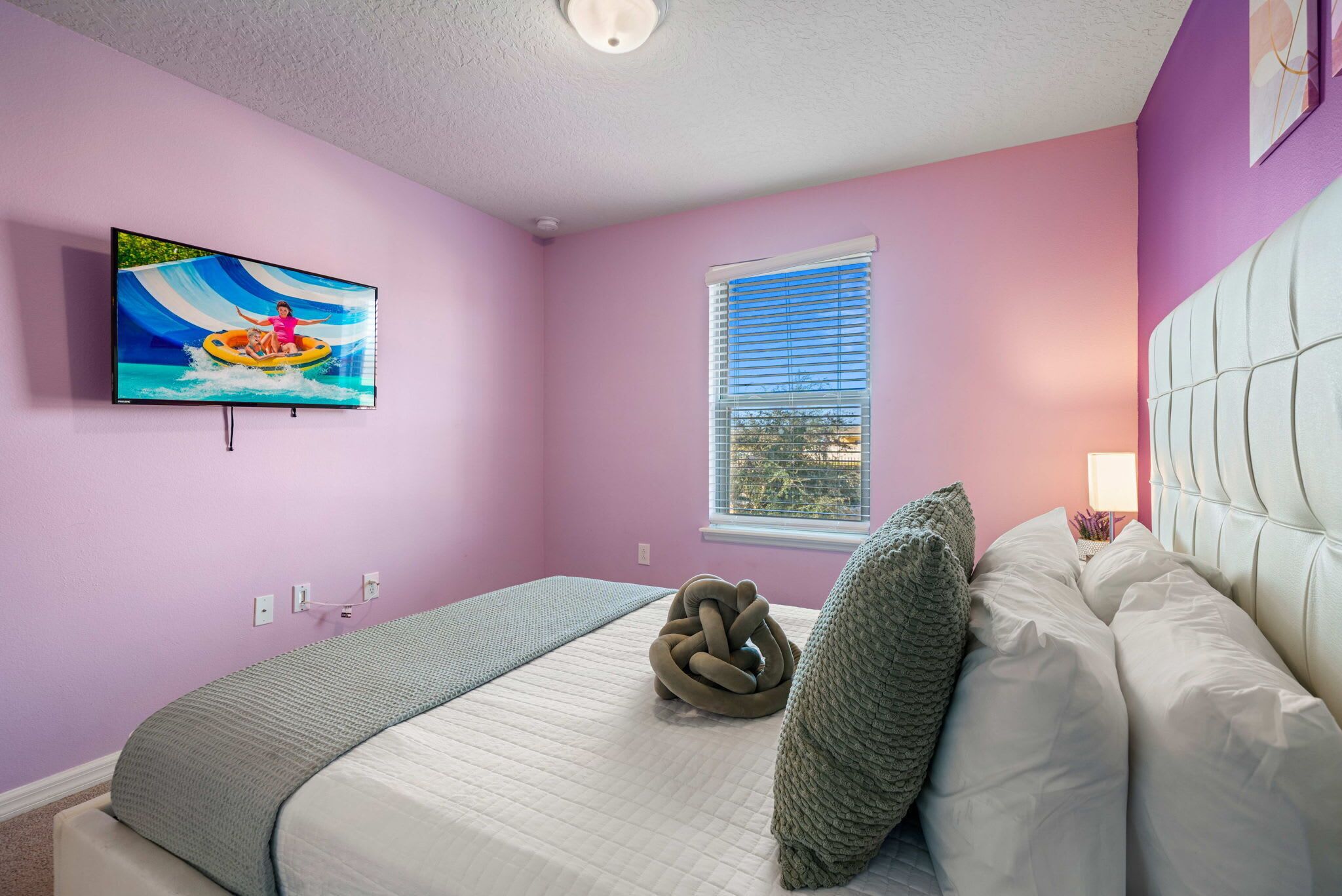 A bedroom with a bed, a TV mounted on the wall, a window, and a view of the outdoors.