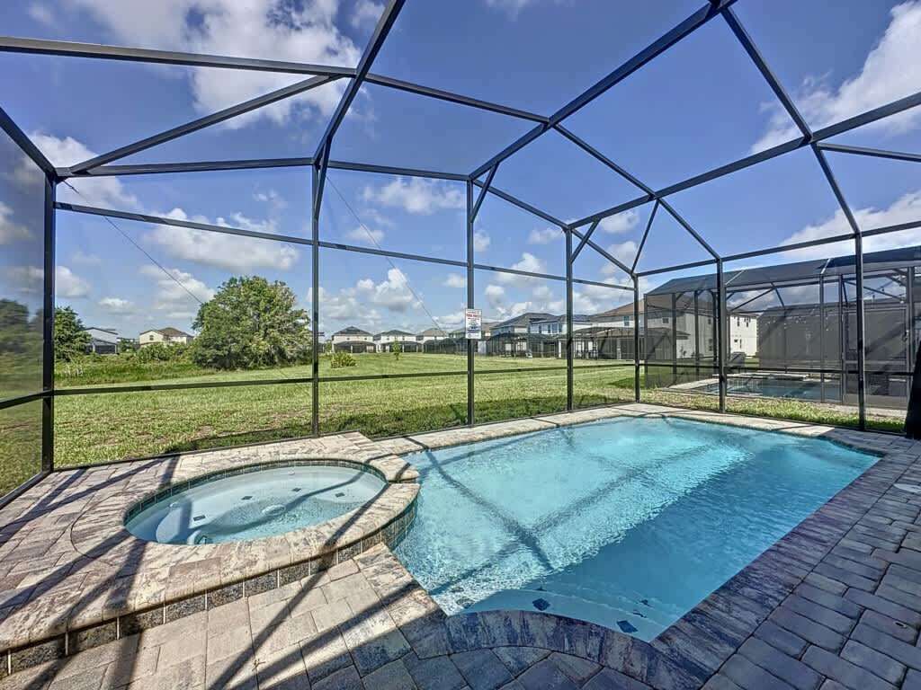 A swimming pool with a hot tub, surrounded by a metal fence, set against a grassy area with houses in the background.
