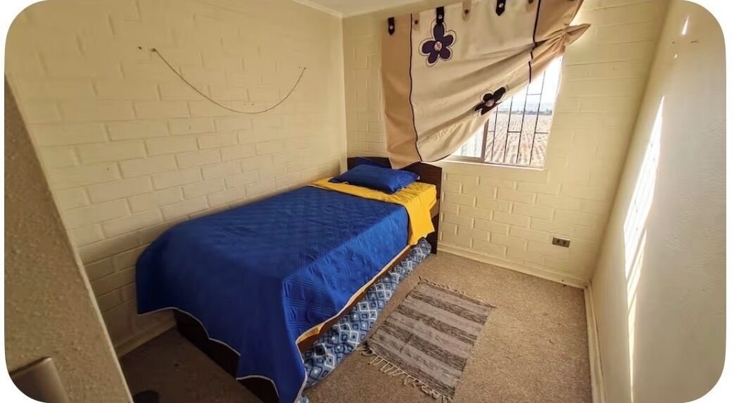 Una piccola camera con un letto singolo, parete con motivo a mattoni, finestra con tende e tappeto decorato.