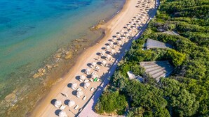 Eine Luftaufnahme eines Strandes mit Reihen von Liegestühlen und Sonnenschirmen, einem klaren, blauen Meer und üppigem Grün auf der rechten Seite.
