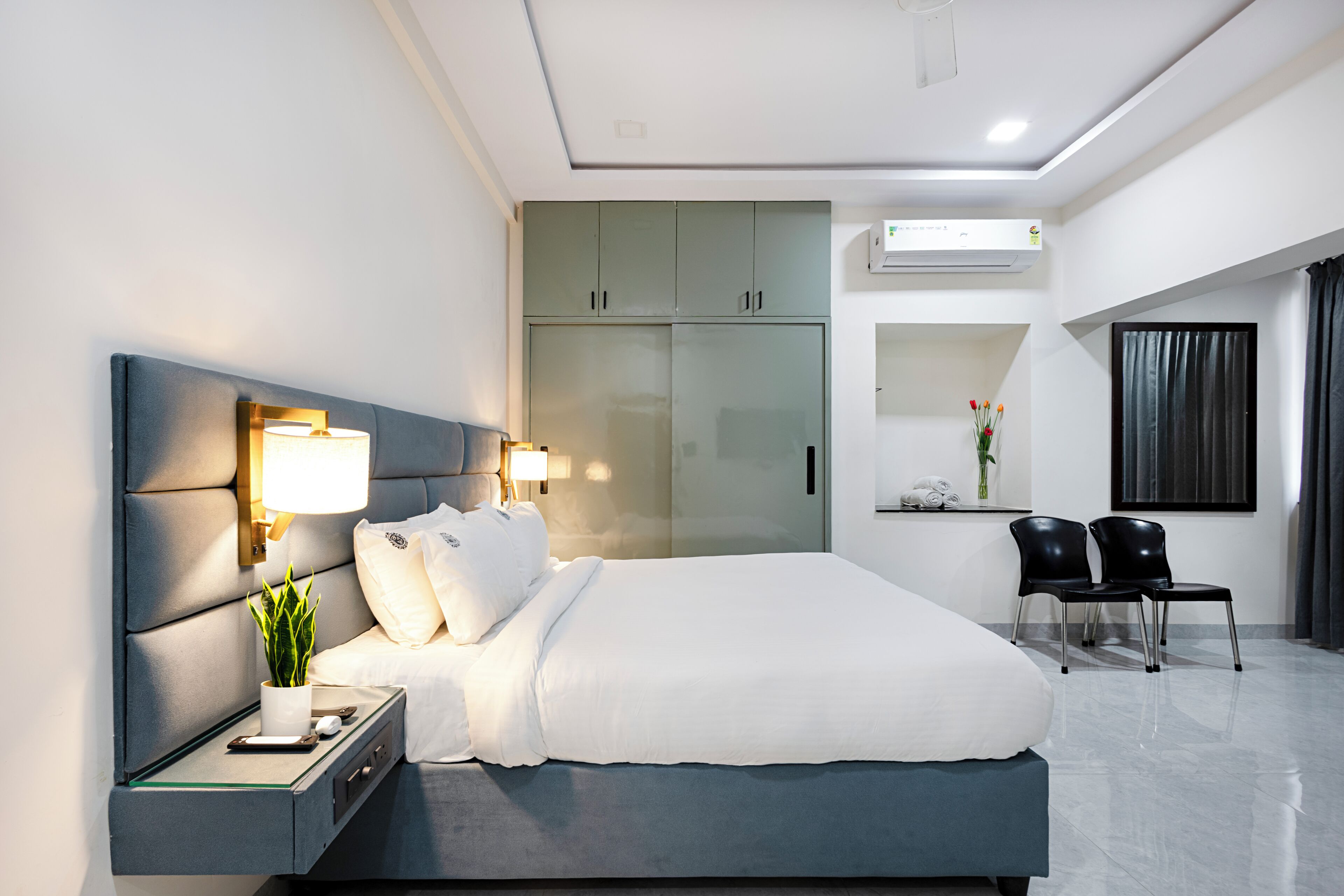 A modern hotel room with a large bed, bedside tables, a desk, a wardrobe, and a seating area with chairs.