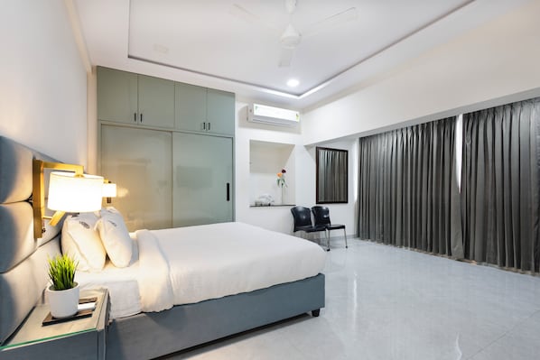 A modern hotel room with a bed, a desk with a lamp, a potted plant, a wardrobe, an air conditioning unit, and a large window with curtains.