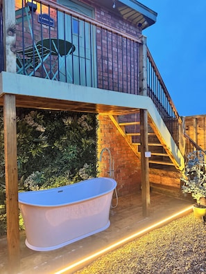 A wooden deck with a large bathtub, a brick wall, and a staircase leading to a balcony.