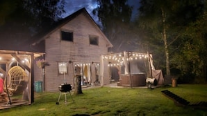 Une maison de campagne en bois, dotée d’un porche couvert et illuminée de guirlandes lumineuses, et entourée d’une pelouse bien entretenue.