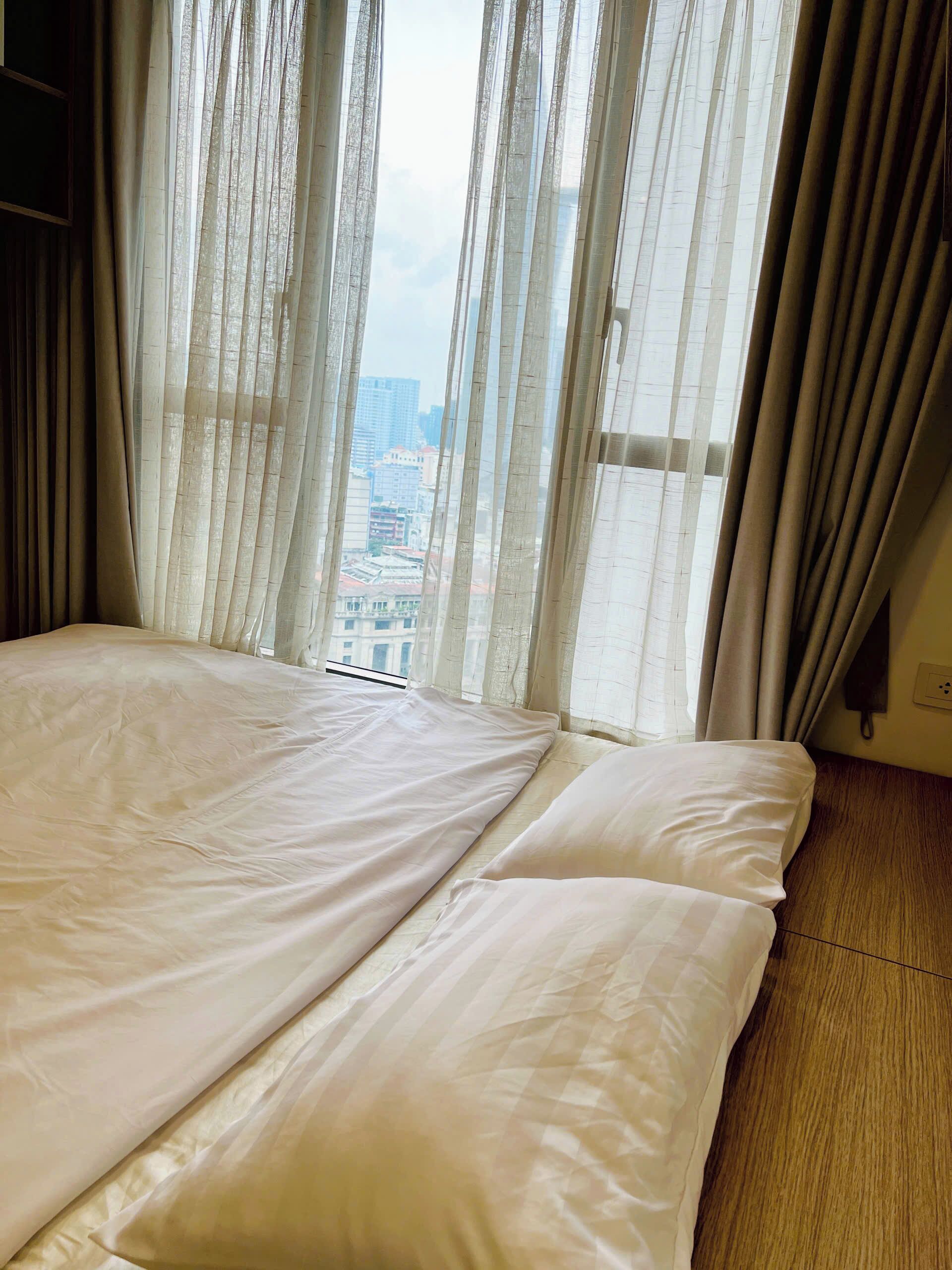 Ein Hotelzimmer mit einem großen Bett, durchsichtigen Vorhängen und Blick auf eine Stadtlandschaft durch das Fenster.