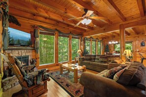 A log cabin interior with a fireplace, wooden furniture, and large windows. - Tennessee Treasure: The Smokies' Best-Kept Secret (Sevierville)