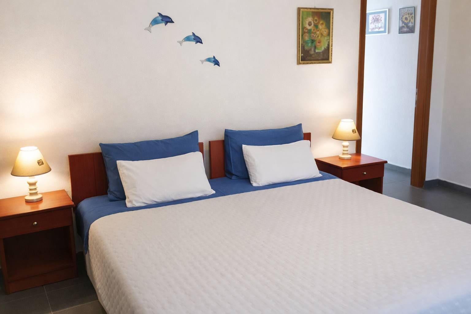 A neatly made bed with blue and white bedding, two bedside tables with lamps, and wall art including a painting and a framed picture.