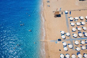 Vista aérea de una playa con aguas azules cristalinas, una orilla arenosa y hileras de sombrillas y sillas de playa.