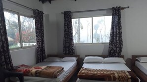 Ein Zimmer mit drei Holzbetten, jedes mit einer anderen Musterdecke. Es gibt Fenster mit Vorhängen, und ein Stuhl ist links im Bild zu sehen.