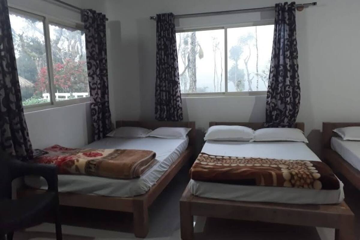 Een kamer met drie houten bedden, elk met een ander beddegoed. Er zijn ramen met gordijnen, en een stoel is aan de linkerkant te zien.