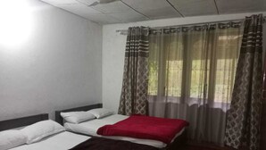 Ein Zimmer mit zwei Betten, eines mit einer roten Decke und das andere mit einer dunklen Decke. Es gibt ein Fenster mit gemusterten Vorhängen.