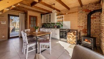 A cozy cabin interior with a wooden dining table, white chairs, a kitchen area with cabinets, and a wood-burning stove.