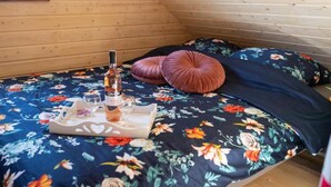Une pièce chaleureuse avec un plafond en pente en bois, un lit recouvert d’une courtepointe à motifs floraux, deux oreillers en forme de cœur et un plateau avec une bouteille et des verres.