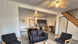 Un salon moderne avec un canapé, deux fauteuils et une table basse. On aperçoit une cuisine au fond.