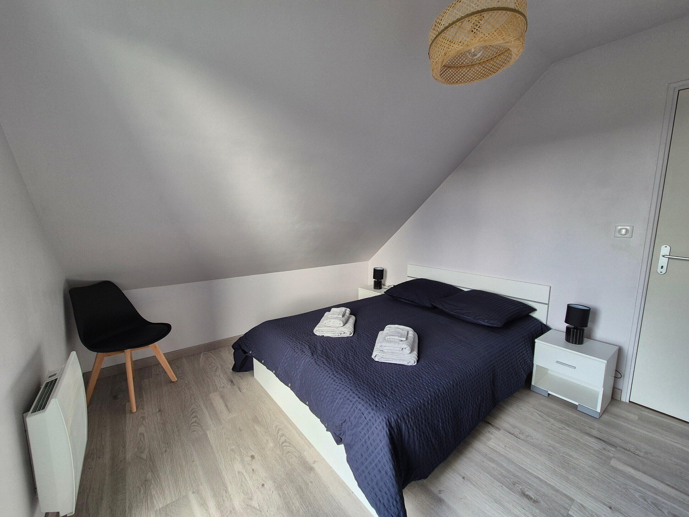 Een slaapkamer met een schuine kap, een bed met donkere beddengoed, een nachtkastje met een lamp en een stoel.