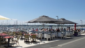 Un espace extérieur aménagé pour les repas, avec des tables et des chaises, une vue sur le port de plaisance en arrière-plan et un ciel bleu dégagé.