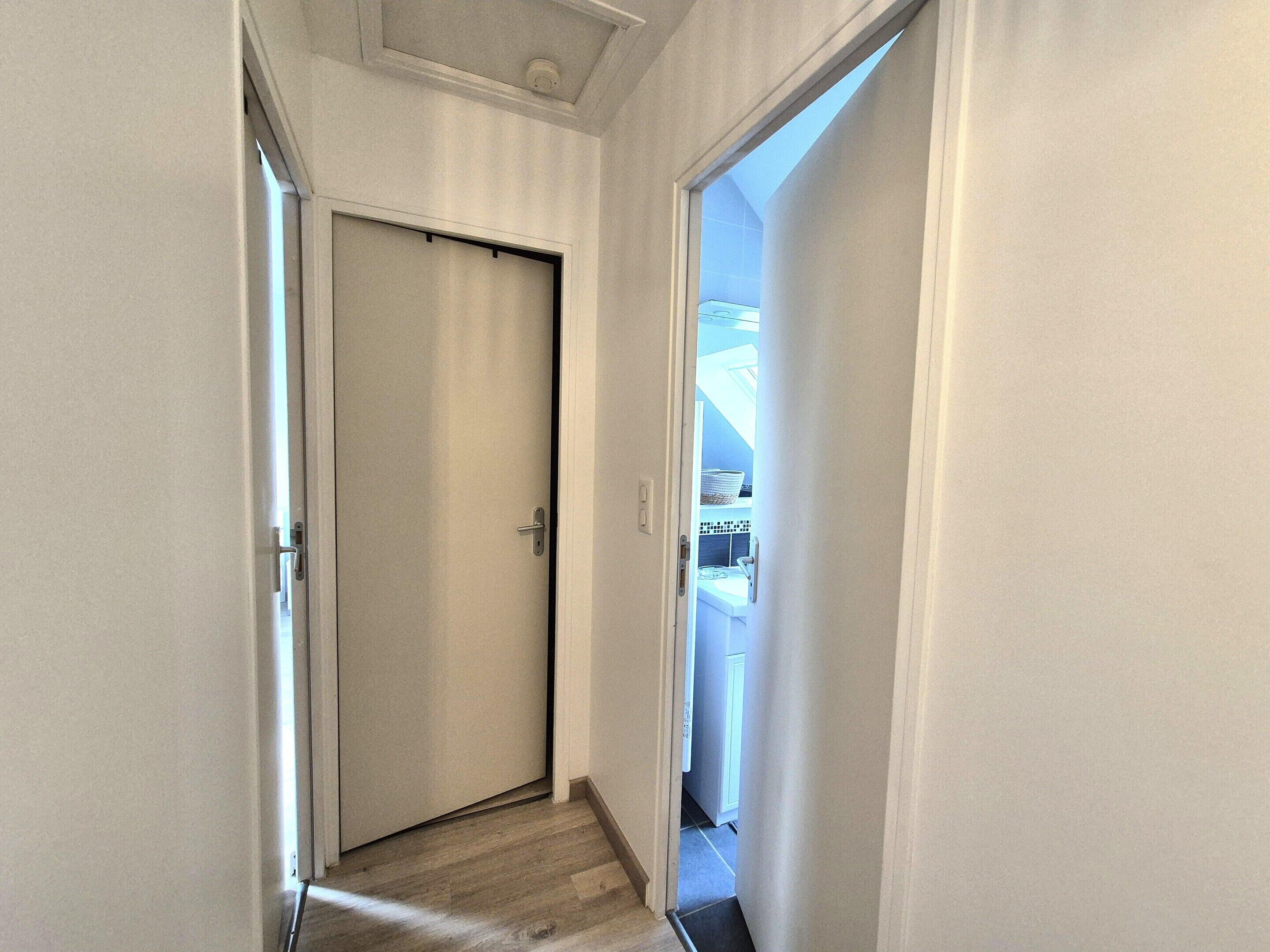 Un couloir étroit, avec une porte fermée au bout, qui donne sur une pièce dont le mur derrière l’évier est recouvert de carreaux et qui possède une fenêtre.