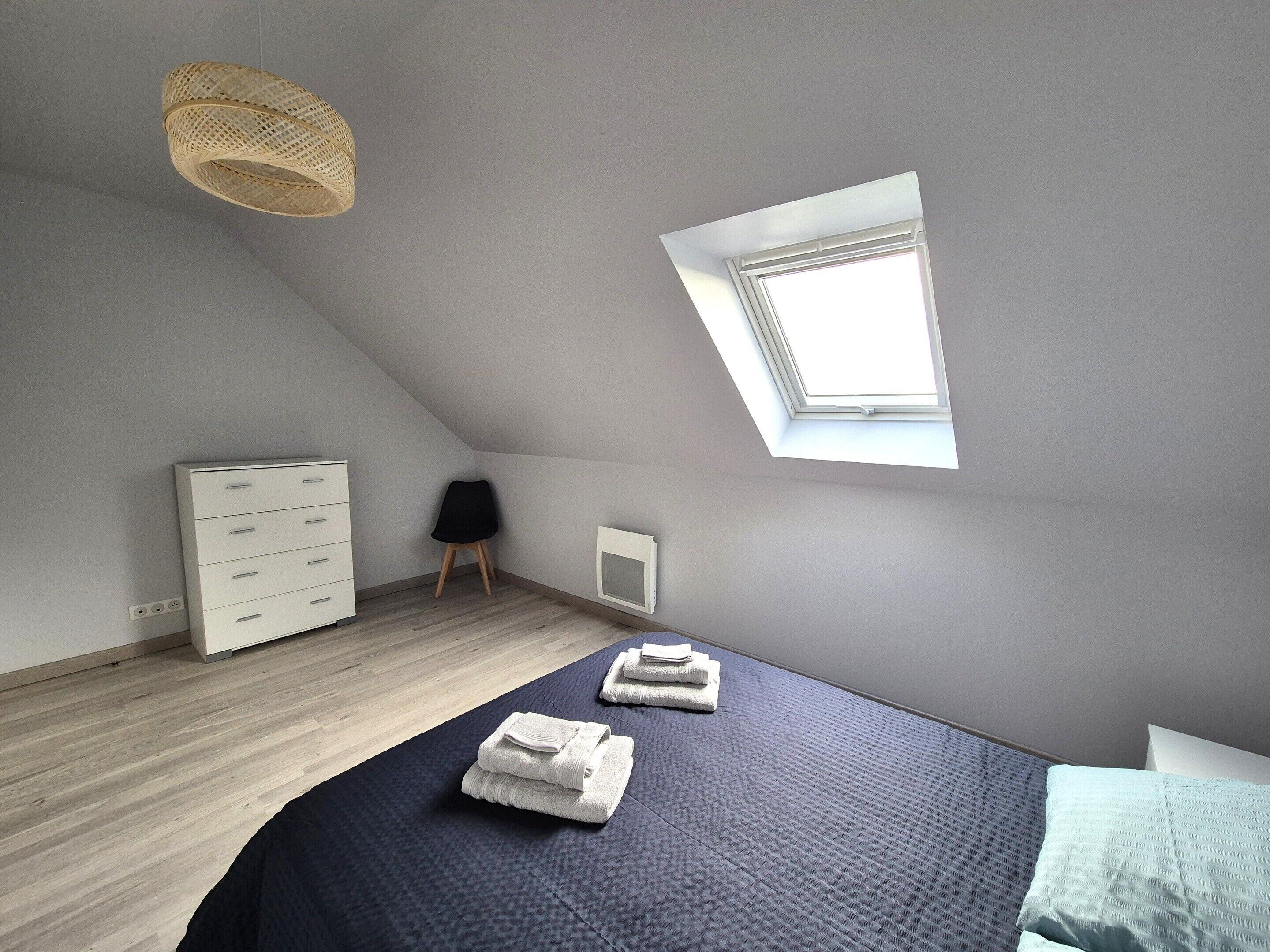 A bedroom with a sloped ceiling, a skylight, a bed with towels, a white dresser, a chair, and a small white cabinet.