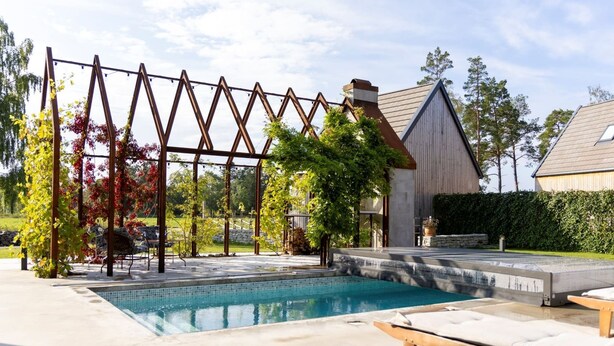 Una piscina con cubierta de madera, sillas de descanso y una estructura de pérgola metálica.
