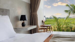A bedroom with a bed, a nightstand with a lamp, a chair, and a view of a green field through a window. - Gangga Homestay Pererenan (Munggu)