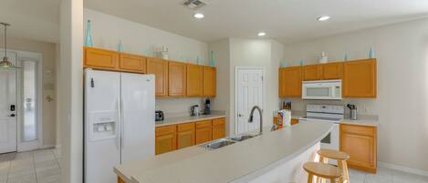 A modern kitchen with wooden cabinets, a white refrigerator, a microwave, and a large island with stools.