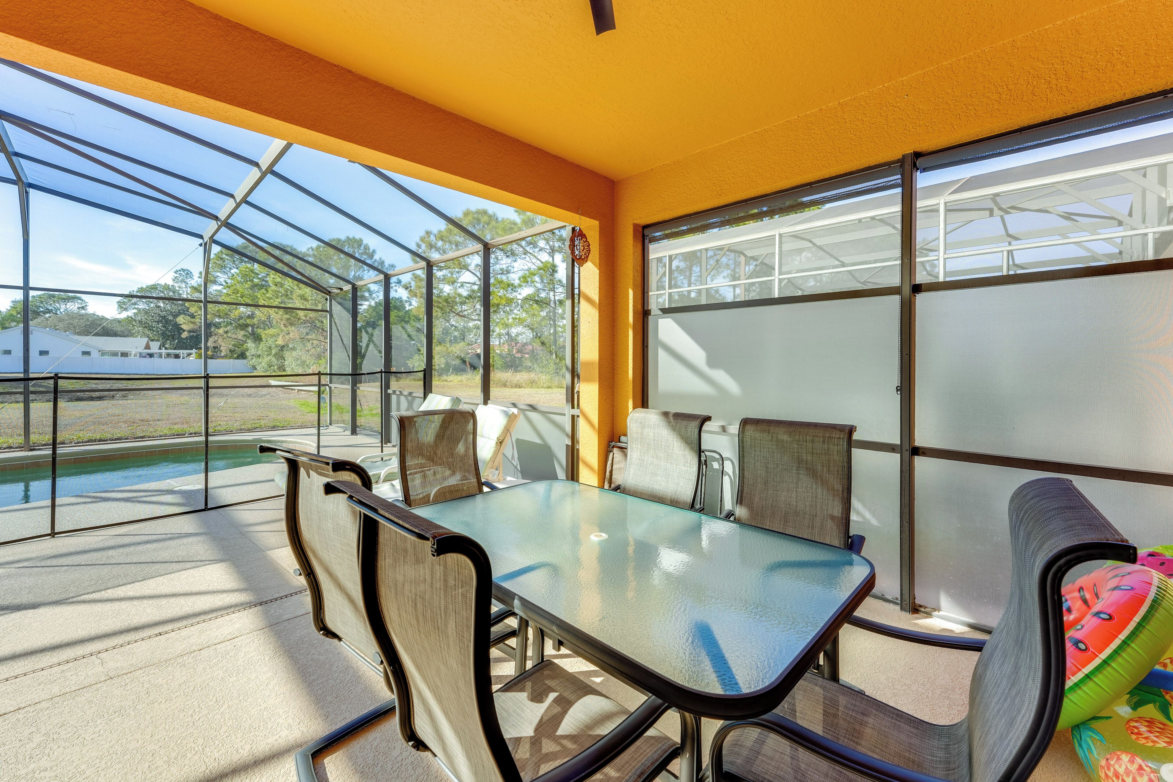 Un espacio de patio techado con una mesa y sillas de vidrio, vista a una piscina y una estructura de invernadero al fondo.
