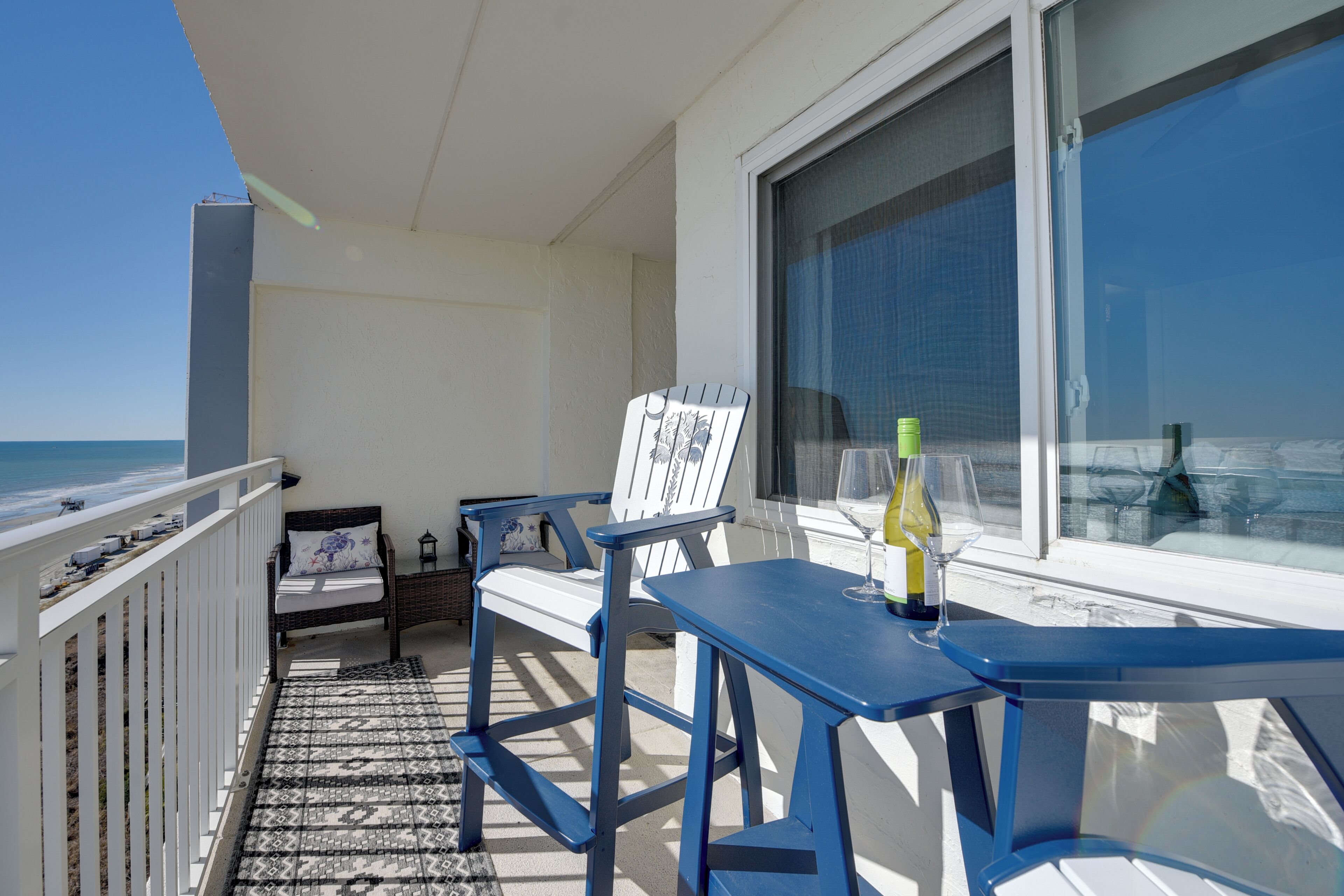 Ein Balkon mit weißer Reling, einem blauen Tisch und einem Stuhl. Auf dem Tisch steht eine Weinflasche.