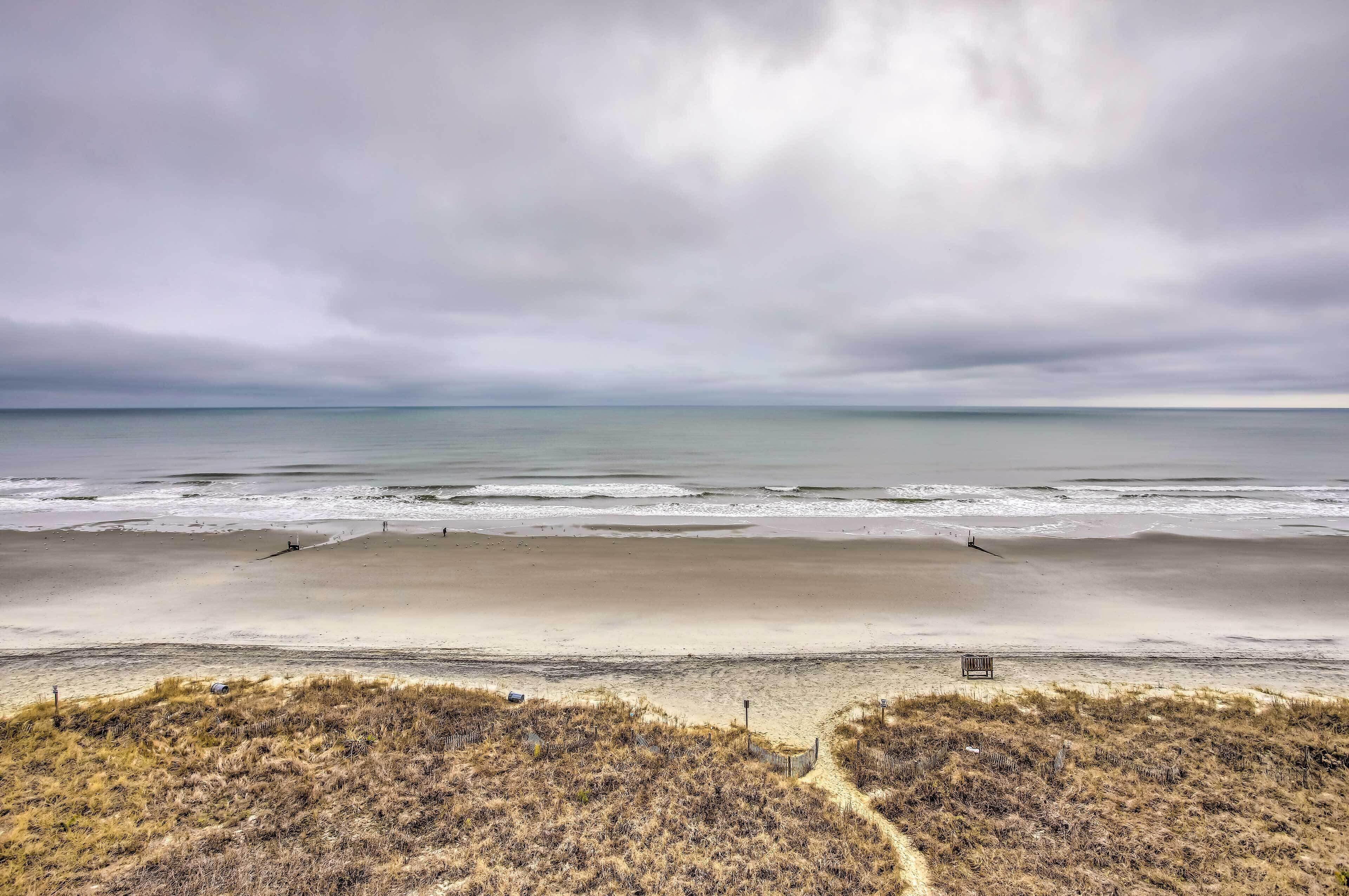 Une plage avec un rivage sablonneux, un tapis d’herbe au premier plan et un ciel nuageux.