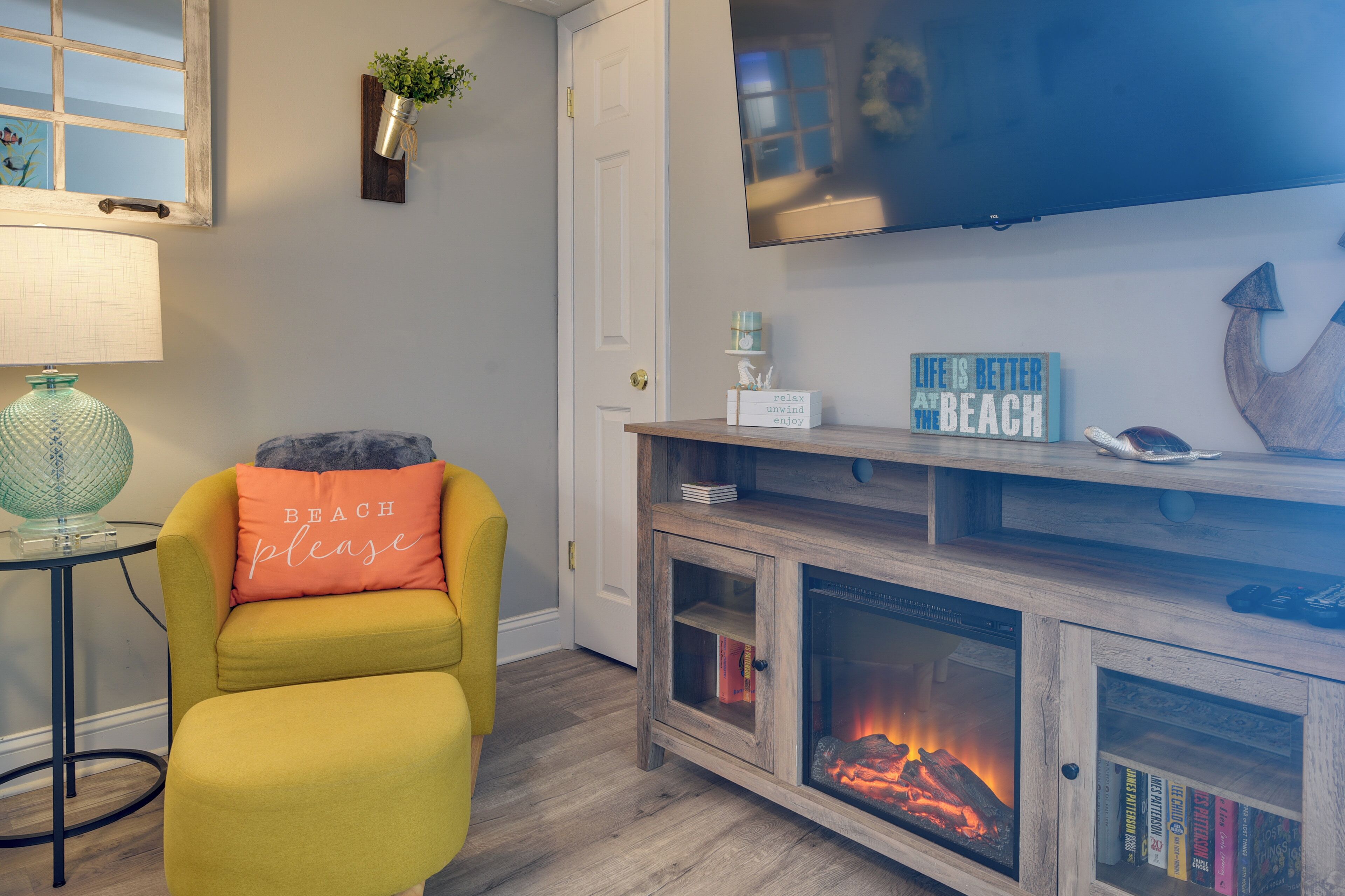A living room with a fireplace, a television mounted on the wall, a yellow armchair, and a small table with a lamp.