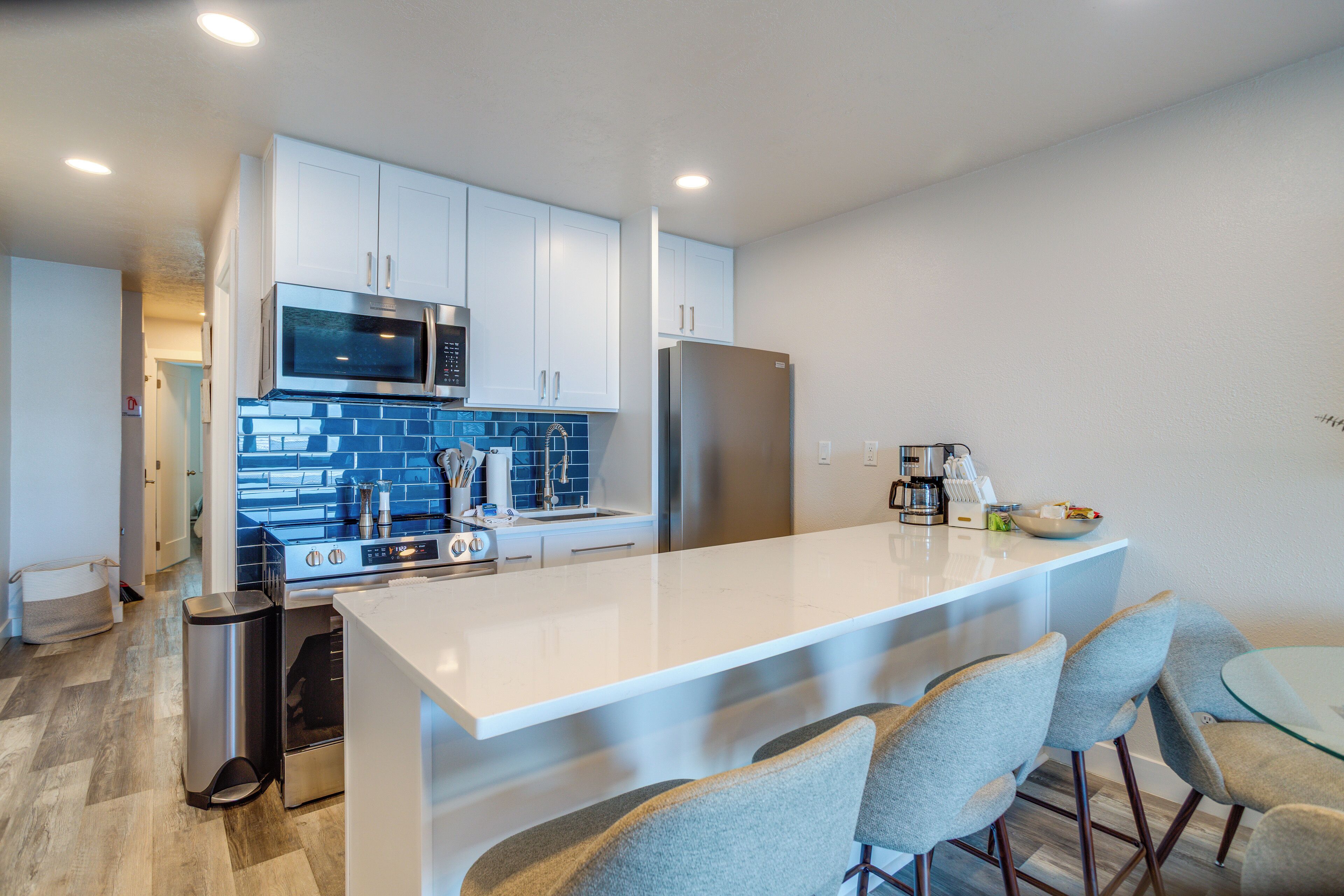 A modern kitchen with white cabinets, a stainless steel refrigerator, a microwave, and a gas stove. There are four bar stools around a central island.