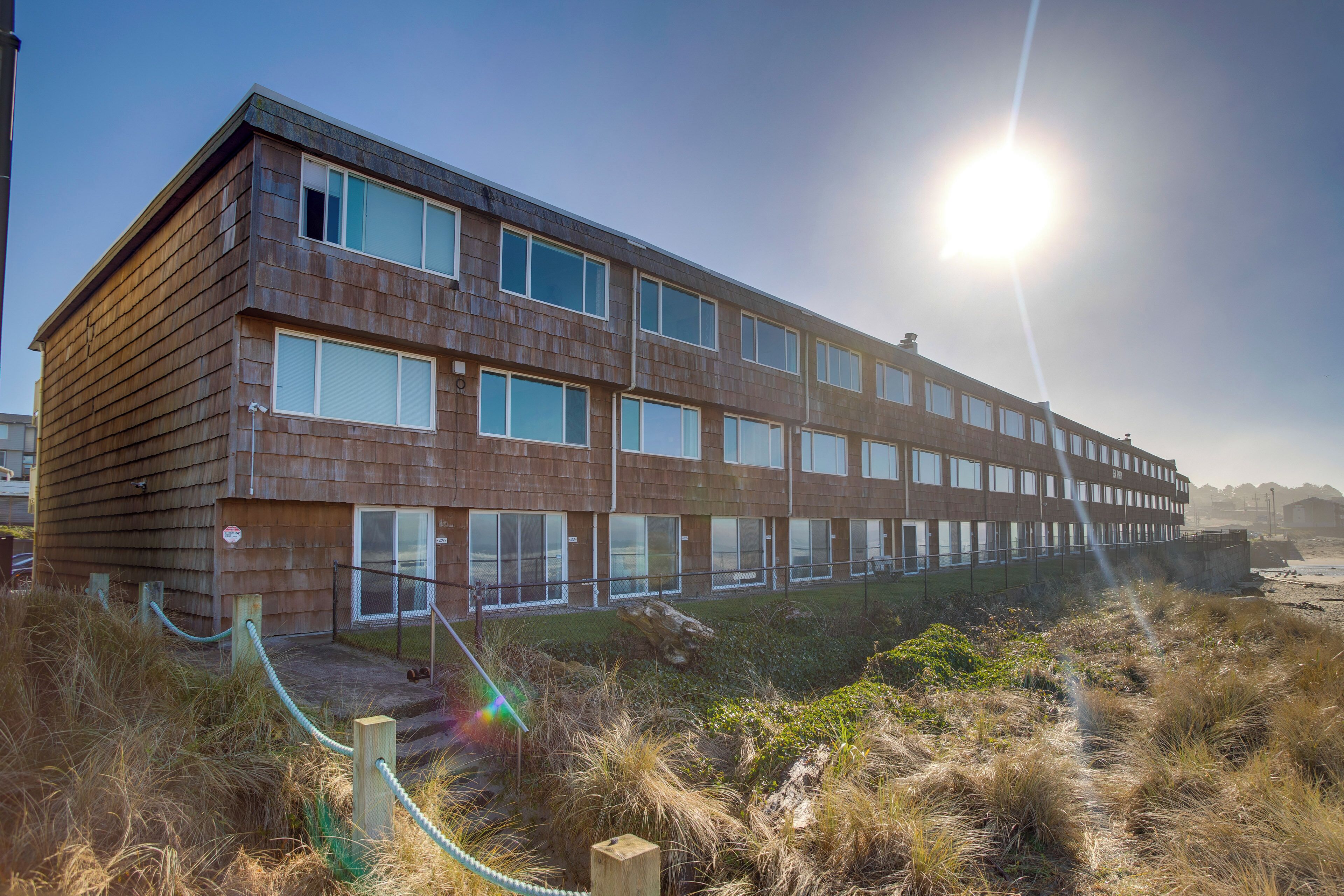 Un bâtiment moderne doté de nombreuses fenêtres et d’une façade en bois, situé près d’une zone verdoyante sous un ciel dégagé.