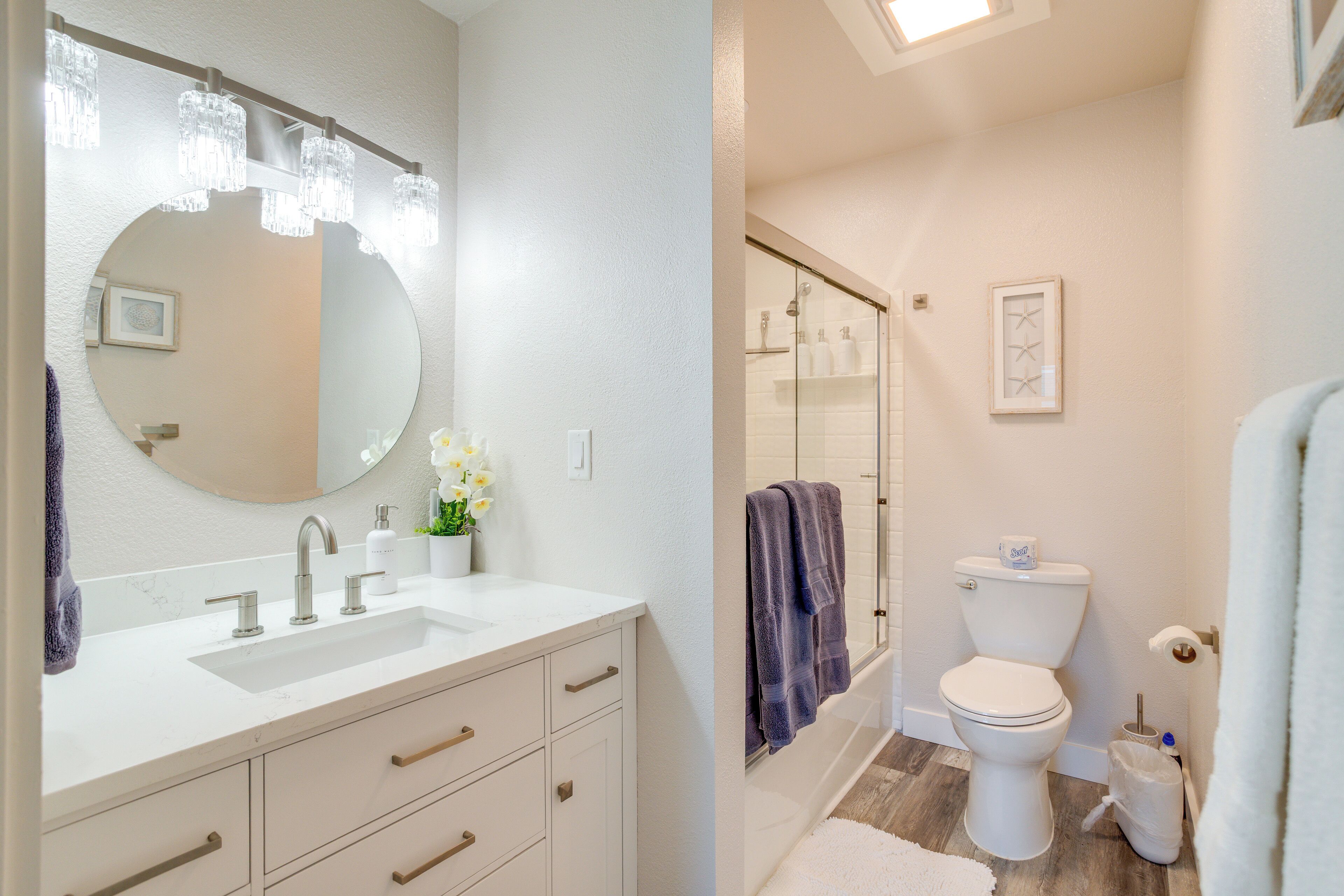 A bathroom with a white sink, mirror, and toilet. There are towels hanging on the wall and a shower area visible.