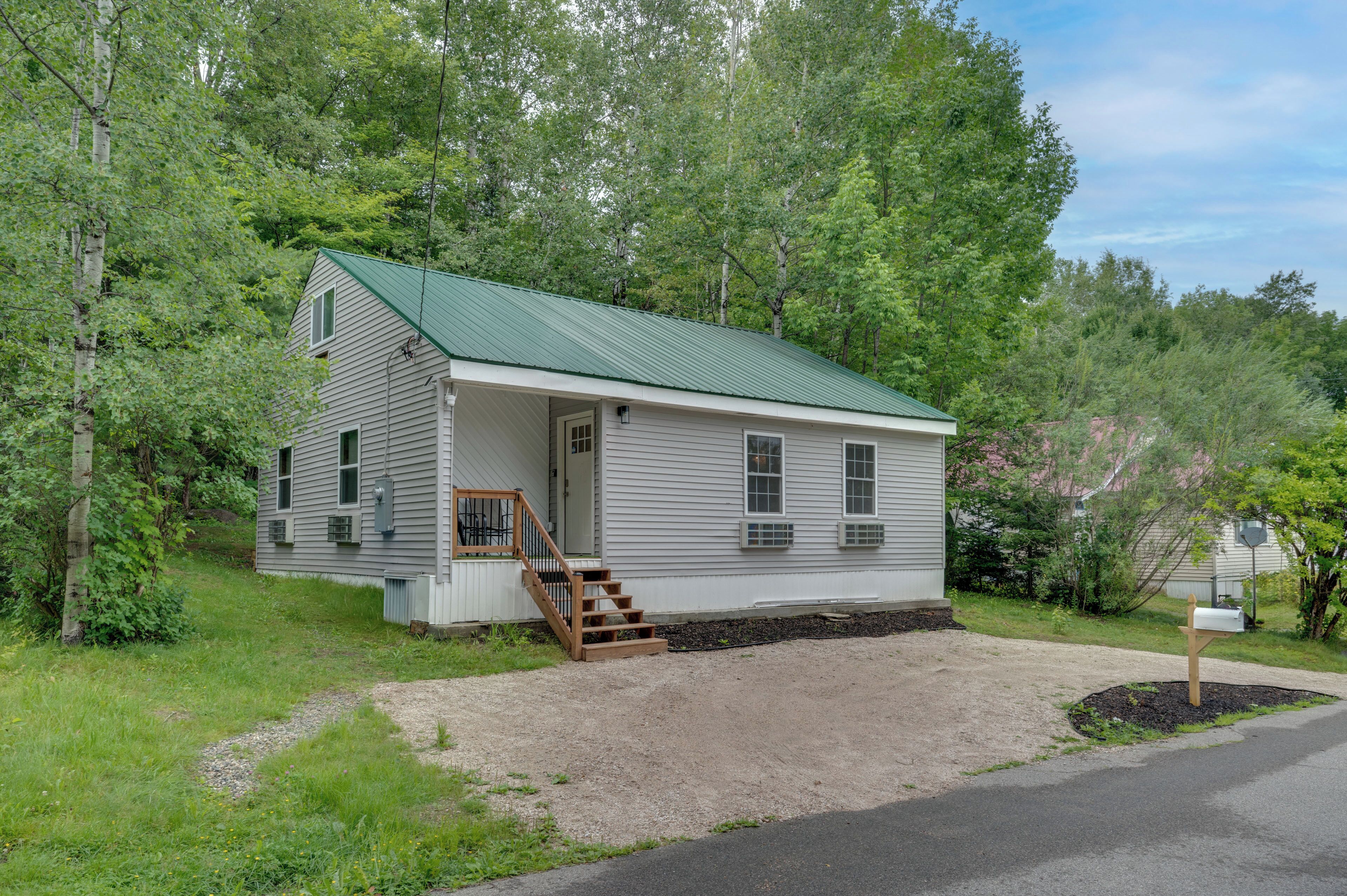 A small, single-story cabin with a green roof, a front porch, and a gravel driveway.
