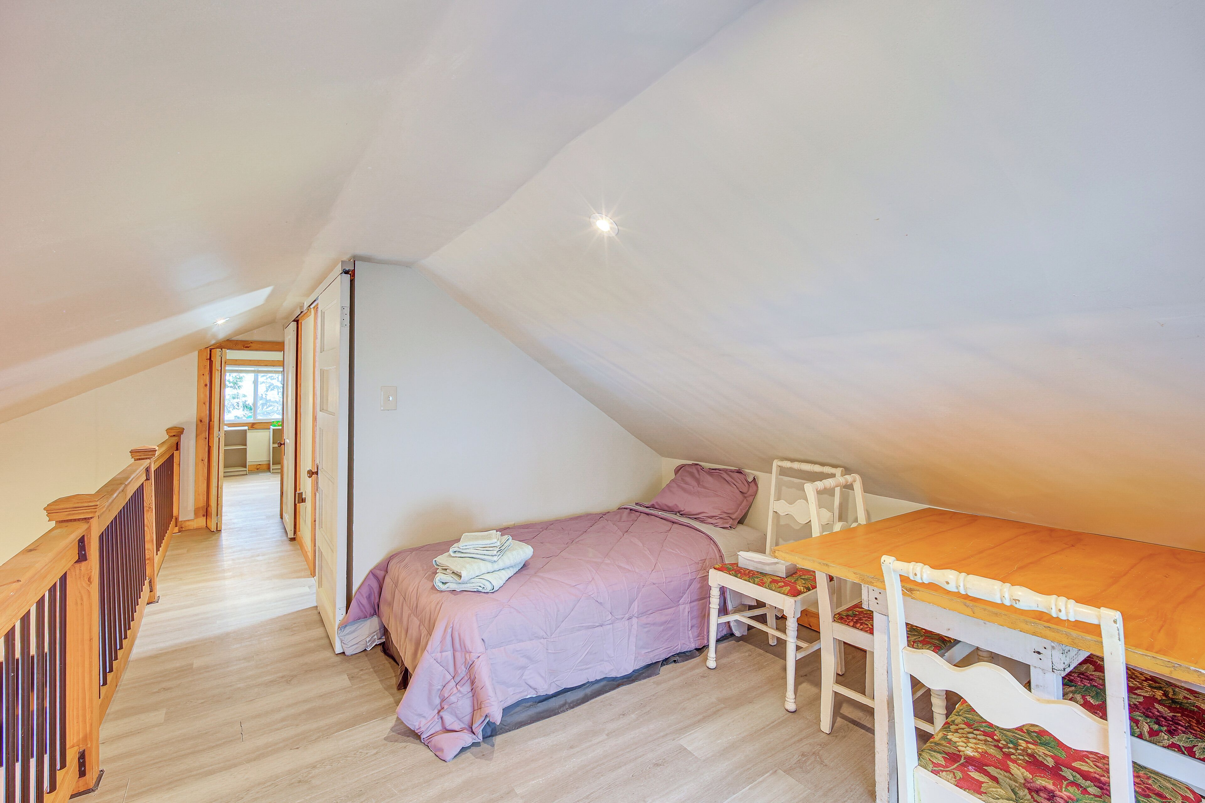 A bedroom with a sloped ceiling, a bed with a pink bedspread, a wooden table with chairs, and a view through an open door.