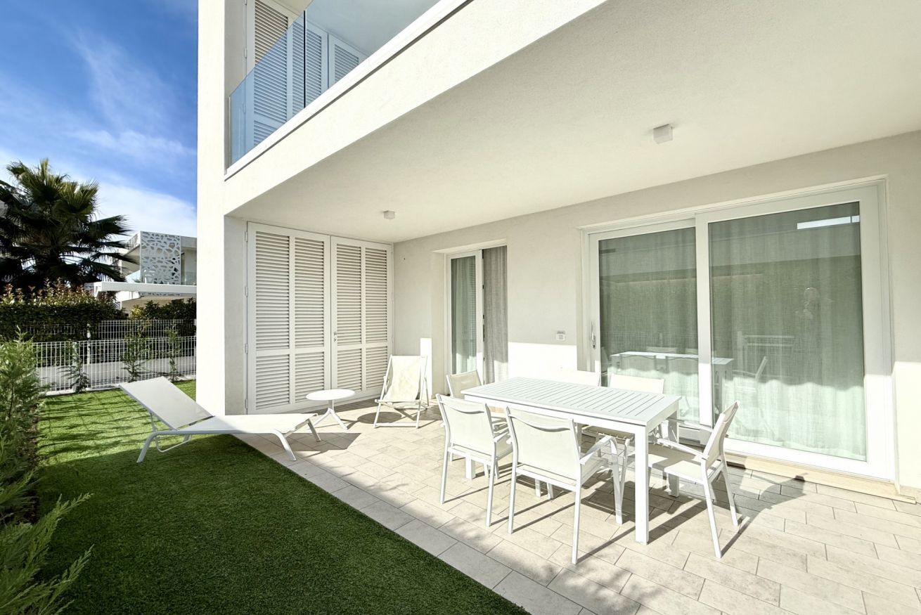 Une terrasse extérieure moderne avec une table et des chaises blanches, une petite pelouse et une porte vitrée donnant sur un balcon.