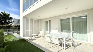 A modern outdoor patio with a white table and chairs, a small lawn, and a glass door leading to a balcony. - Le Jolie 1 - Carraro Immobiliare Jesolo (Jesolo)