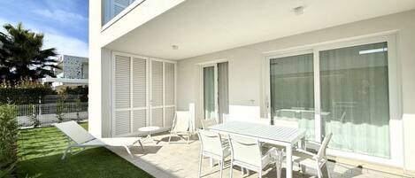 Une terrasse extérieure moderne avec une table et des chaises blanches, une petite pelouse et une porte vitrée donnant sur un balcon.