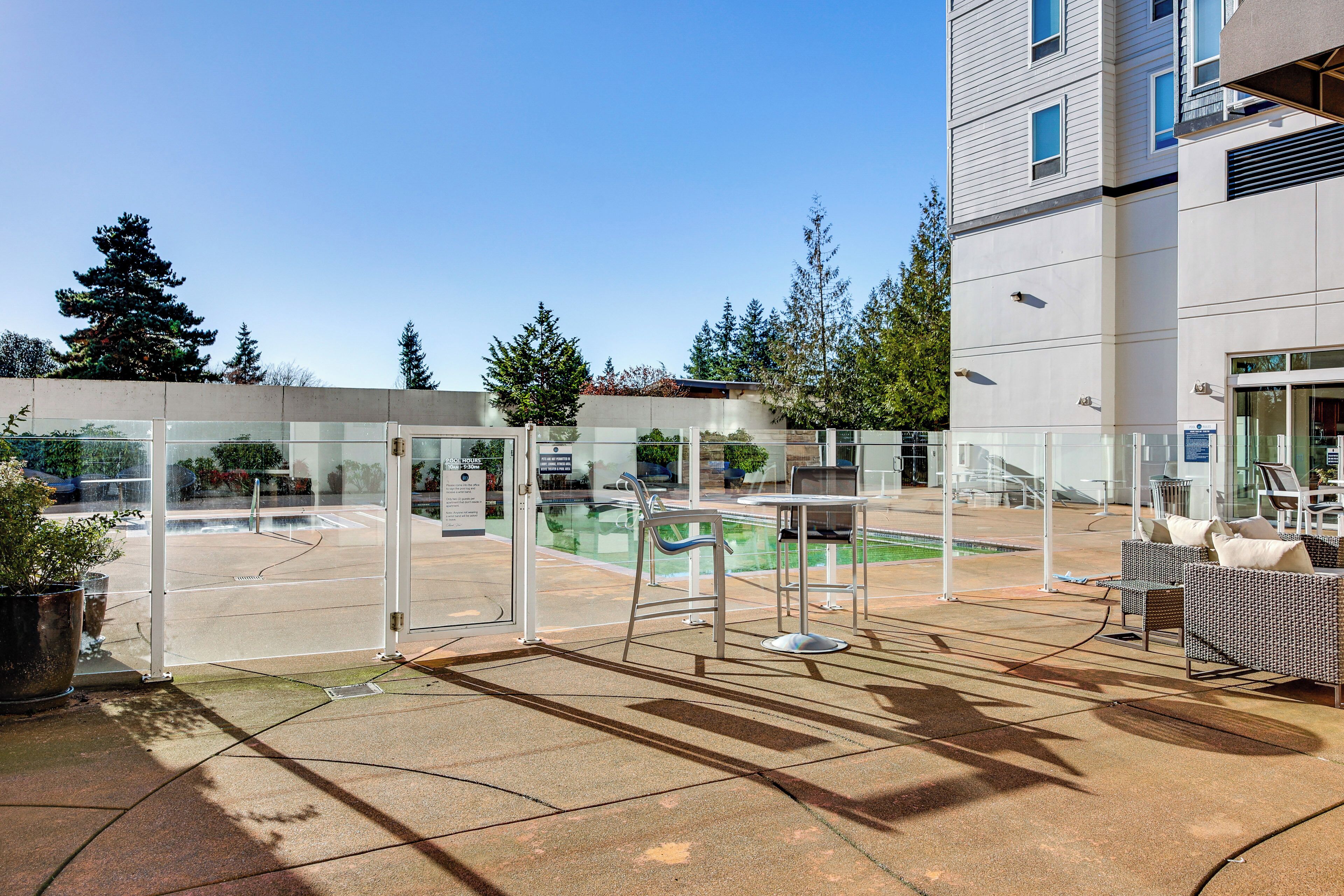 A pool area with glass fencing, outdoor seating, and a building in the background.