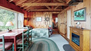 Ein gemütliches Blockhaus-Interieur mit Holzbalkendecke, Kamin, Essbereich mit Tisch und Stühlen sowie einem Bett mit karierter Tagesdecke.