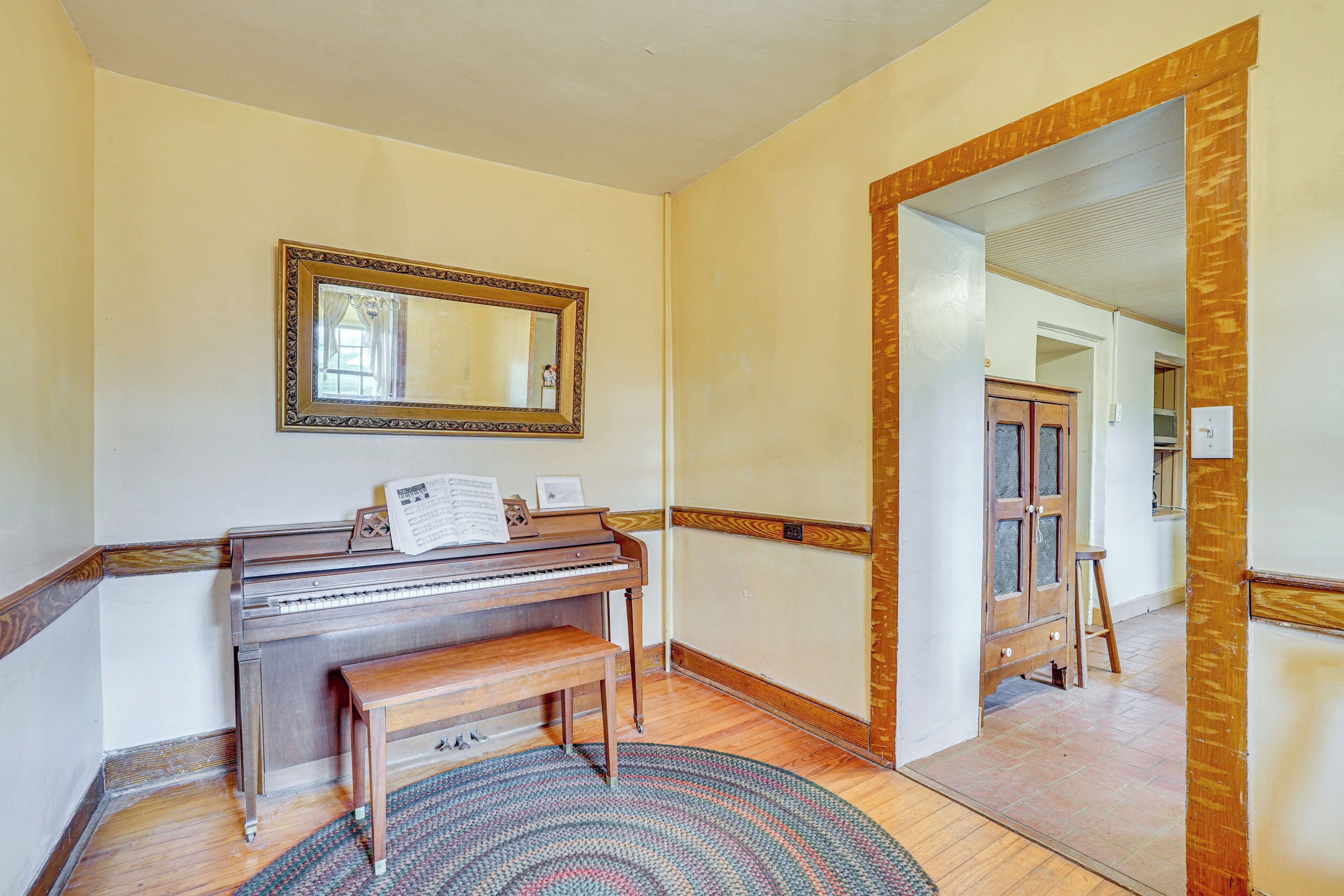 A piano with sheet music, a mirror, and a rug in a room with wooden floors and a wooden door frame.