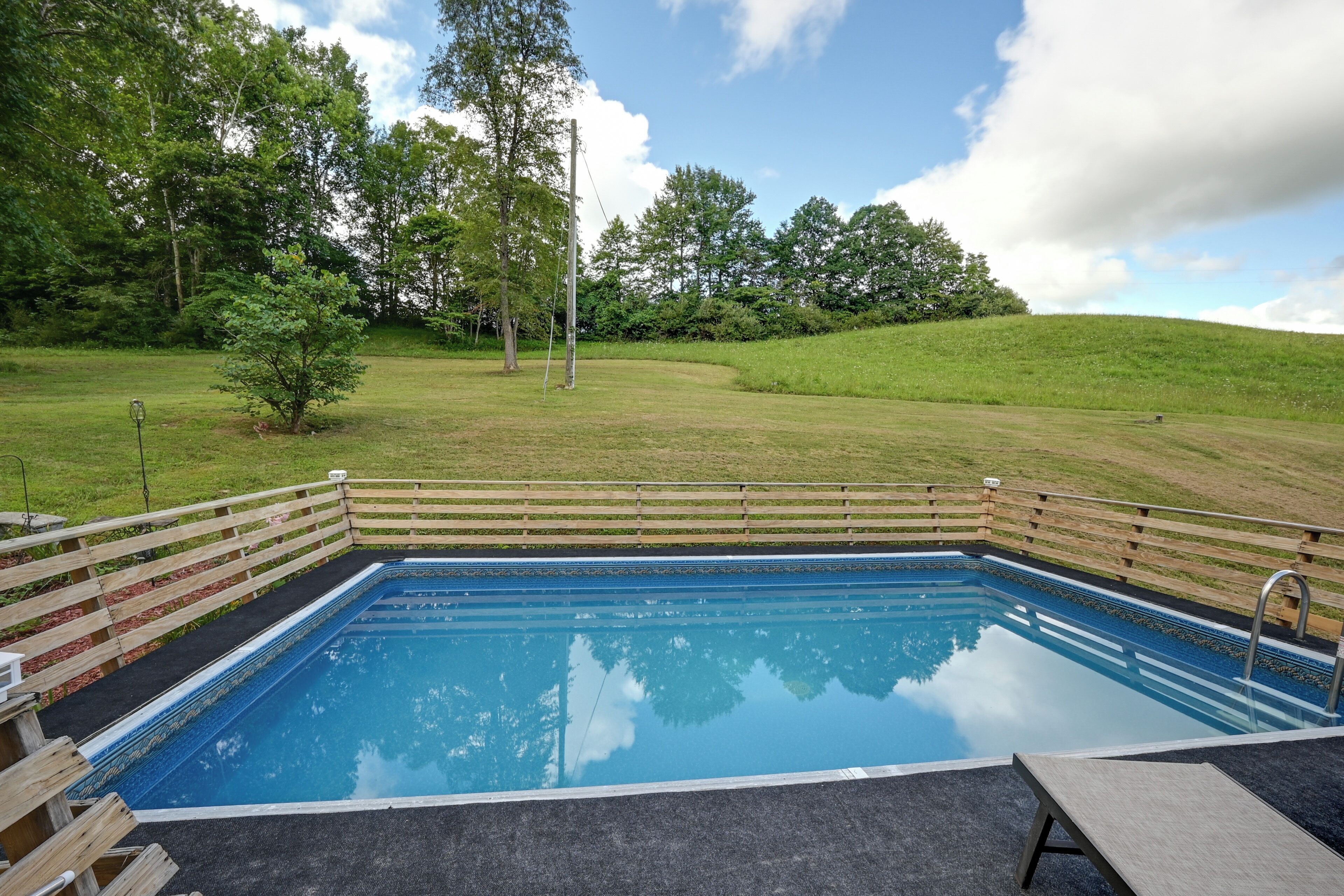 A rectangular swimming pool with a wooden deck surrounding it, set against a backdrop of a grassy hill and trees.