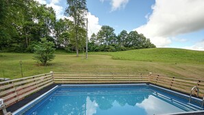 Ein rechteckiger Pool mit umlaufender Holzterrasse, vor einer sanften Hügelkuppe mit Bäumen.