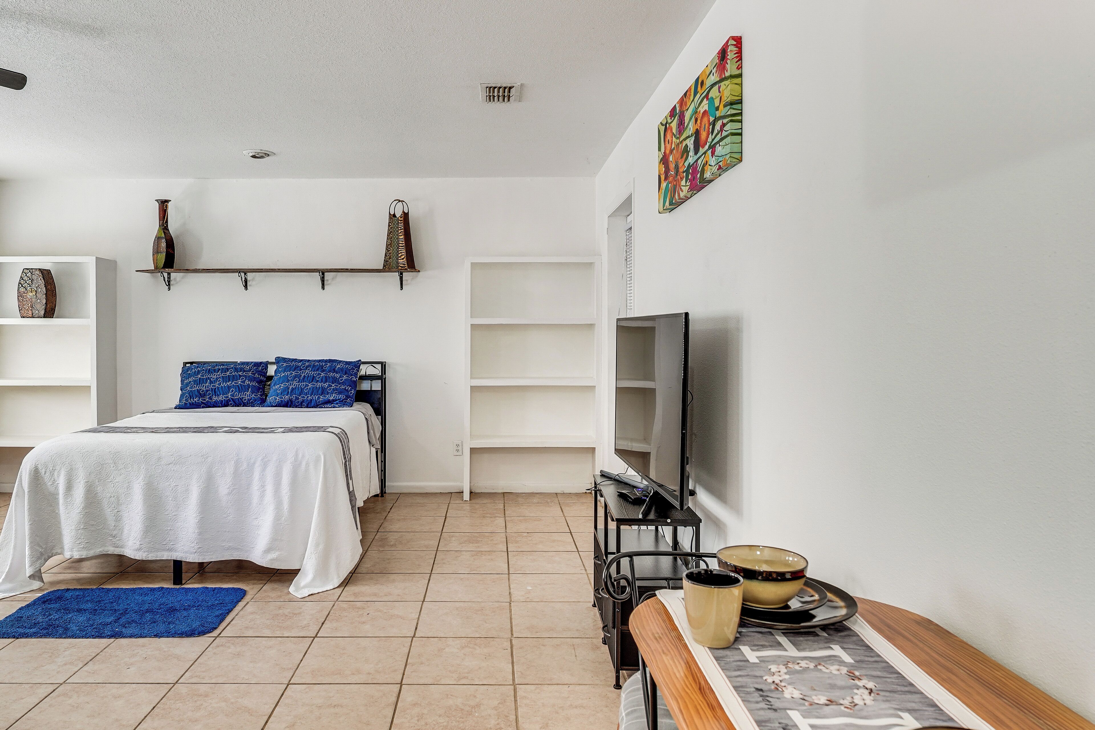 Une chambre à coucher avec un lit, une télévision, une étagère ornée d’objets décoratifs et un tapis bleu.