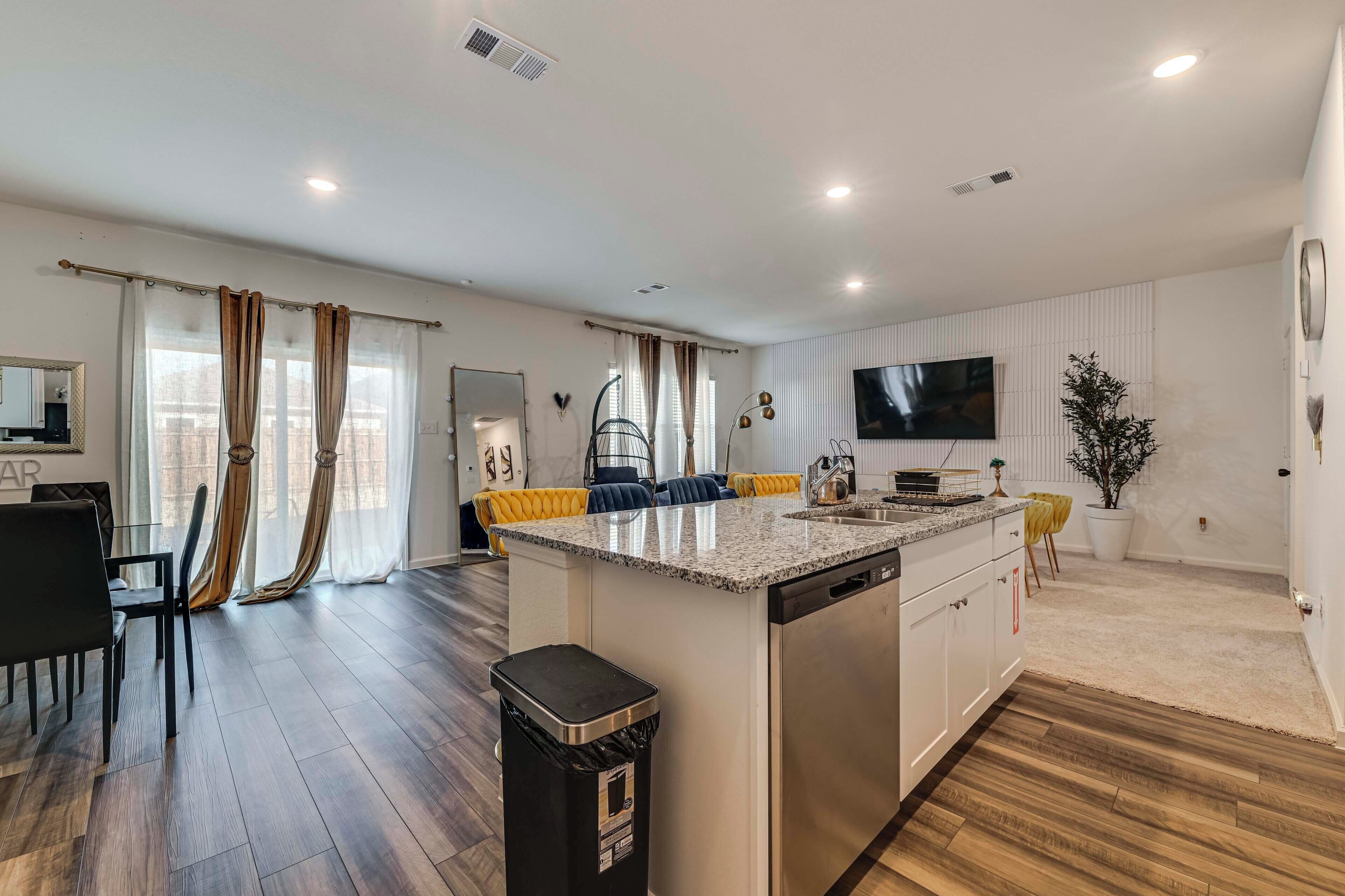 A modern kitchen with a central island, a dining area with a black chair, and a living space with a TV and curtains.