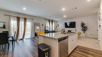A modern kitchen with a central island, a dining area with a black chair, and a living space with a TV and curtains.