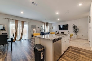 A modern kitchen with a central island, a dining area with a black chair, and a living space with a TV and curtains. - Group Haven in Princeton! WFH Ready & Ample Space (Princeton)