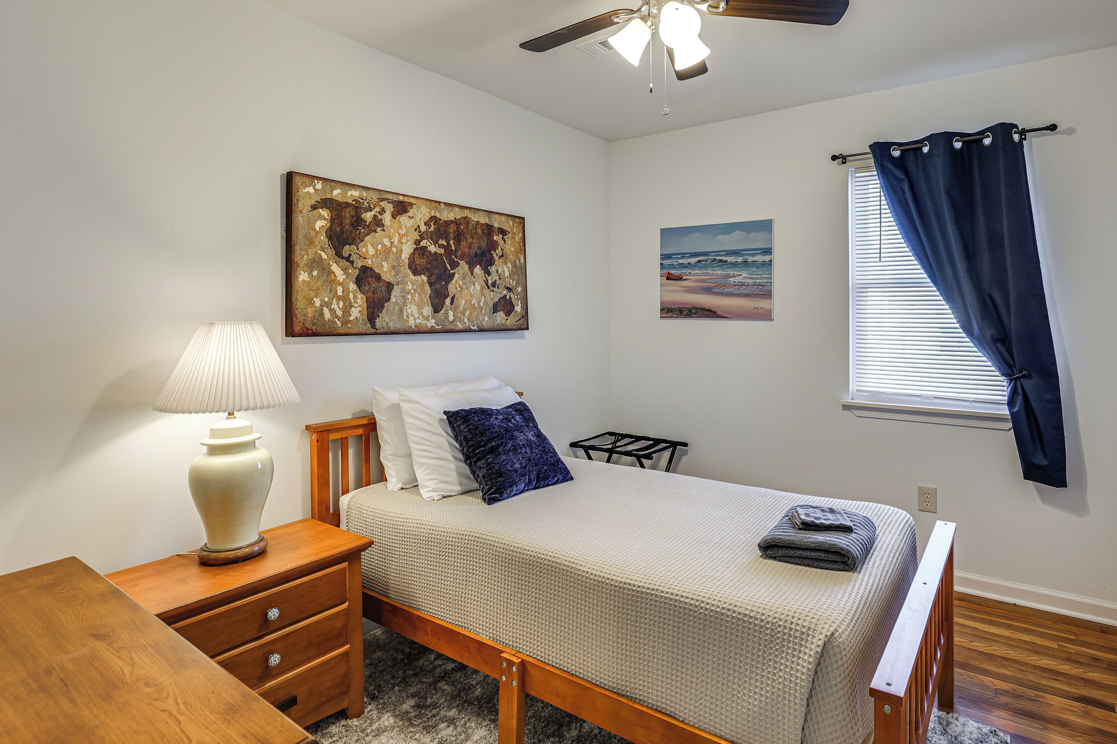 A bedroom with a bed, a nightstand, a lamp, a ceiling fan, a window with curtains, and artwork on the walls.